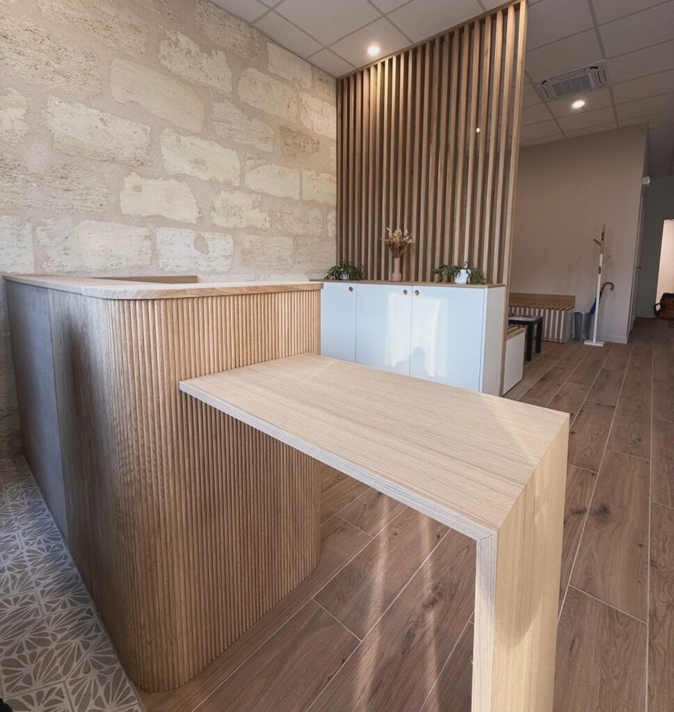 Accueil moderne : Comptoir bois, design à lattes et mur pierre. Comptoir d'accueil moderne en bois clair à lattes verticales, devant un mur en pierre naturelle. Sol carrelé, cloison en bois et meuble blanc avec plantes.