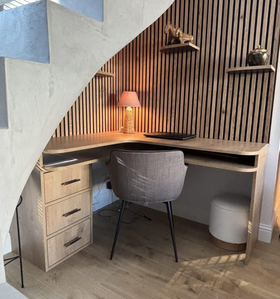 Bureau sous escalier : Aménagement élégant et fonctionnel Bureau d'angle en bois clair sous escalier moderne. Mur à lattes de bois, chaise grise, lampe chaleureuse et étagères décoratives.