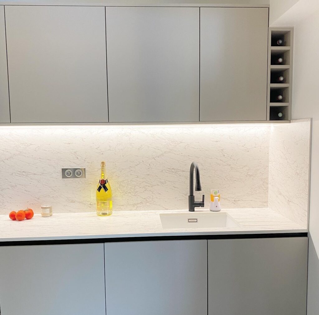 Cuisine moderne élégante : armoires grises, marbre et éclairage LED. Cuisine moderne aux armoires grises, comptoir et crédence en marbre clair. Éclairage LED, évier, robinet noir, casier à vin. Bouteille Moët & Chandon et tomates.