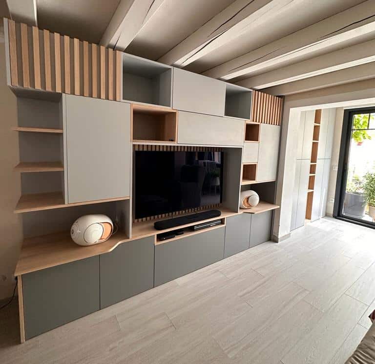 Meuble TV mural design sur mesure | Rangement moderne salon Grand meuble TV mural sur mesure gris et bois clair avec niches, étagères, caissons bas gris foncé. Deux enceintes design blanches. Plafond à poutres.