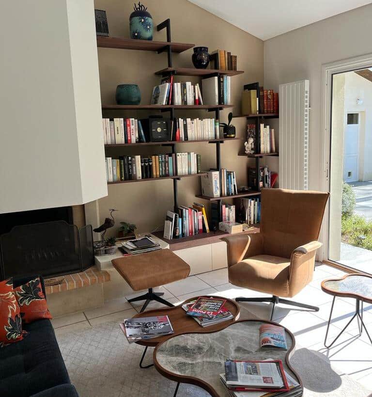 Salon Moderne Cosy : Bibliothèque Murale Design & Fauteuil Salon moderne avec grande bibliothèque murale, fauteuil marron, pouf, tables basses et cheminée. Ambiance lumineuse et cosy.