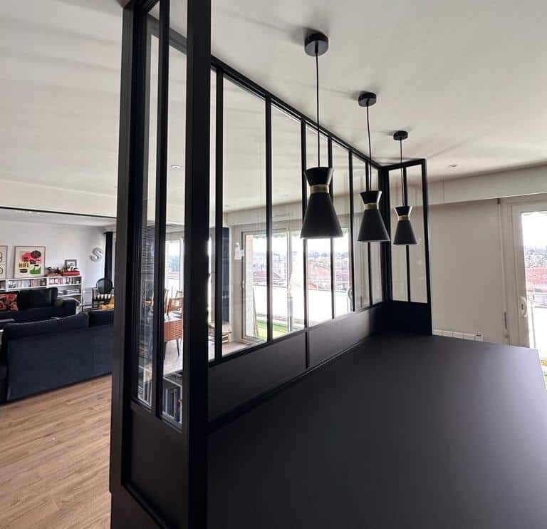 Verrière Noire Moderne : Design Salon et Cuisine Ouverte Intérieur moderne avec verrière noire et vitrée séparant un salon lumineux (sol bois, canapé, HIFI) d'un comptoir sombre sous trois suspensions dorées. Vue sur la ville.